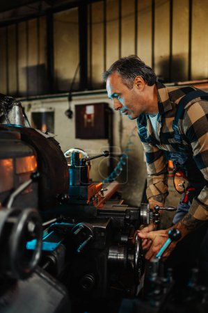 Photo d'un machiniste expérimenté mesurant et fixant une vieille machine à tour dans un atelier vintage. Il est concentré et professionnel, immergé dans le processus. Il porte une chemise à carreaux et des salopettes.