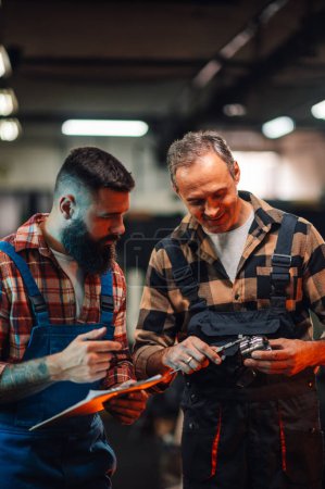 Two metallurgy engineers checking the metrics of a cylinder steel element in a factory. The grey haired man is holding a caliper and a metal bearing, the bearded young man is holding a clipboard.