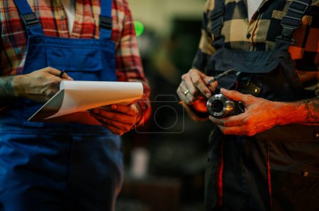 Close up of hands of two metal turning workers. One person is holding a caliper and a metal fitting and the other is holding a clipboard and a pen. Hands holding cylinder and clipboard.