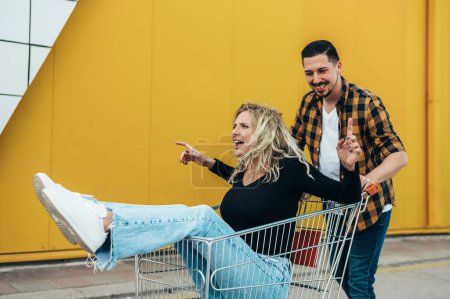 Jeune beau couple chevauchant dans un panier de supermarché et s'amusant devant un fond jaune