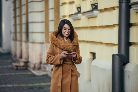 Jeune femme hispanique utilisant un smartphone alors que dans la ville portant un manteau