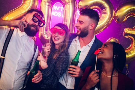 Beautiful young diverse friends having fun at New Year's Eve Party in the club. Two couple having fun while celebrating together with alcohol drinks and party photo props. Multiracial friends.