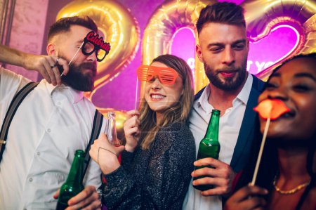 Young multiracial friends having fun at New Year's party while drinking alcohol and dancing. Posing with funny photo props. Party, holidays, celebration, nightlife and people concept. Copy space.
