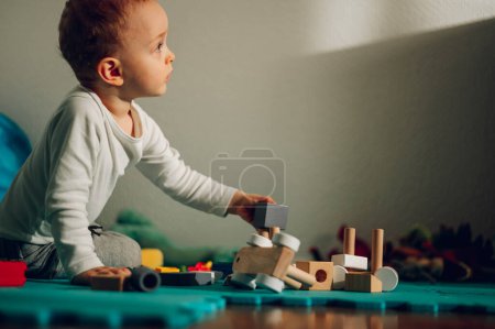 A curious adorable child is sitting on the floor at home and learning shapes through the game with a wooden Montessori toy. Learning new skills, imagination, and healthy development.