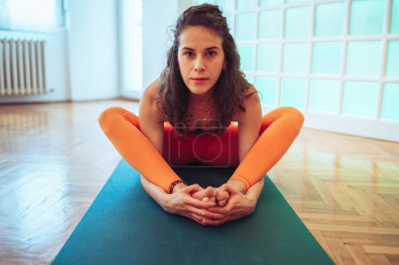 Wide shot of a young curly caucasian girl sitting on a yoga mat and doing butterfly exercise while stretching legs before splits. Looking straight to the camera while leaning forward. Copy space.