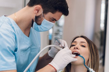 Männlicher Zahnarzt fixiert Zähne einer Patientin im Zahnarztstuhl. Zahnarzt, der die Zähne der Patienten untersucht und sich in der Zahnklinik untersuchen lässt. Arzt mit elektrischem Handstück oder Luftturbine.