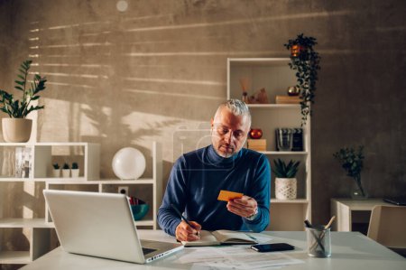 Gray haired middle man aged using credit card and a laptop while sitting in a home office. Gray haired man using online banking and paying bills and rent with an online banking. Copy space.