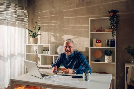 Gray haired middle aged man holding credit card and looking into the camera while sitting in a home office. Smiling guy online shopping and paying with an online banking. Copy space.