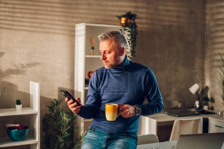 Hombre guapo de mediana edad sentado en una mesa en una oficina en casa y usando un teléfono inteligente mientras bebe café. El tipo de pelo gris escribiendo un mensaje en un teléfono móvil y bebiendo café de la mañana. Copiar espacio.
