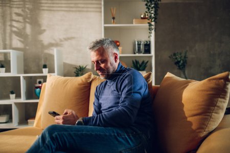 Portrait of a smiling grey hair man with beard sitting on a couch at home and using a smartphone. Middle aged guy in casual clothes typing on a mobile phone and spending free time at home. Copy space.