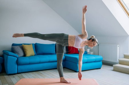 Jeune belle femme formation yoga à la maison un debout dans l'exercice équilibre asana. Tuladandasana pose. Entraînement à domicile avec débardeur et leggings.