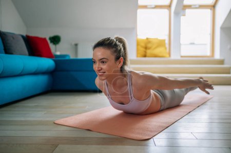 Belle femme souriante pratiquant le yoga à la maison et faisant de l'exercice Double Leg Kicks tout en étant allongé sur un tapis de fitness. Salabhasana pose. Yoga à la maison.