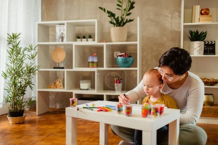 Porträt einer Mutter und ihres kleinen Sohnes beim gemeinsamen Spielen und Zeichnen am kleinen Tisch zu Hause. Babysitterin oder Tante, die dem kleinen Jungen hilft und dabei Spaß hat. Glückliche Familie. Kopierraum.
