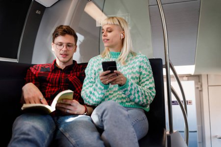 Jeune couple heureux à conduire dans un métro ou un train et passer leur temps ensemble. Amis joyeux souriant et parlant ensemble tout en voyageant ensemble tout en lisant un livre et en utilisant un téléphone.