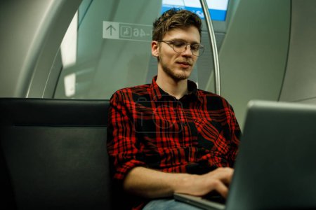 Portrait d'un jeune hipster monté dans un train ou un métro et utilisant un ordinateur portable pendant son travail ou ses études. La vie quotidienne et les trajets domicile-travail ou une école par les transports publics. Espace de copie.