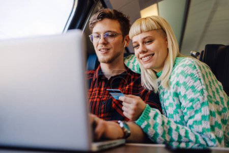 Retrato de una feliz pareja de viajeros que viajan en tren y utilizan la tarjeta de crédito para reservar en línea en el portátil. Jóvenes pasajeros viajando en tren y sonriendo a la computadora portátil mientras compran en línea.