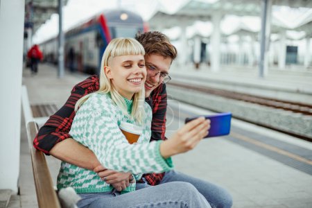 Portrait d'un jeune passager branché assis sur une gare, embrassant et faisant des selfies tout en souriant au téléphone. Les navetteurs attendent un train à la gare et prennent des autoportraits.