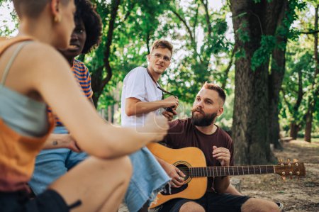 Group of interracial group of environmentalists sitting in a park with guitar and discussing ecological issues and global warming. Group of eco friends promoting ecology and earth day in nature.