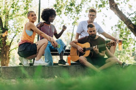 Eine Gruppe von vier jungen künstlerischen Freunden hängt an einem sonnigen Tag im Park herum, spielt Gitarre und genießt rhythmische Musik. Vier urbane junge Erwachsene genießen freien Nachmittag in der Natur und musizieren.