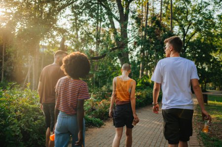 Gruppe multiethnischer Freunde, die eine Pause vom urbanen Lebensstil einlegen und einen Spaziergang in einem Park genießen, während sie ihre Getränke trinken. Rückansicht von Freunden, die gemeinsam spazieren gehen und die Natur erkunden.
