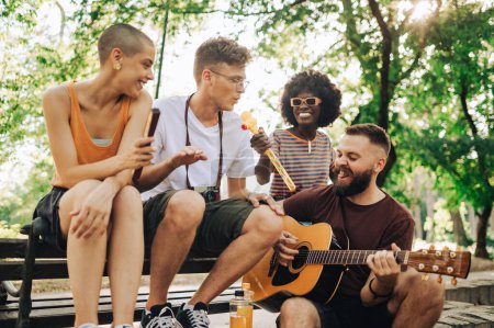 Eine kleine lustige Gruppe von Freunden sitzt im Stadtpark und spielt Gitarre und singt. Wenige multikulturelle Freunde amüsieren sich im Stadtpark. Ein junger Mann spielt Gitarre und singt draußen.