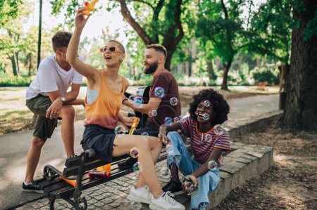 Eine kleine Gruppe multikultureller Freunde sitzt im Stadtpark und amüsiert sich. Ein Mädchen mit kurzen Haaren spielt mit Blasen und macht Stimmung, während ein junger Mann im Hintergrund eine Gitarre spielt.