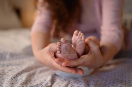 Mother holding tiny baby feet in her hands while on the bed at home