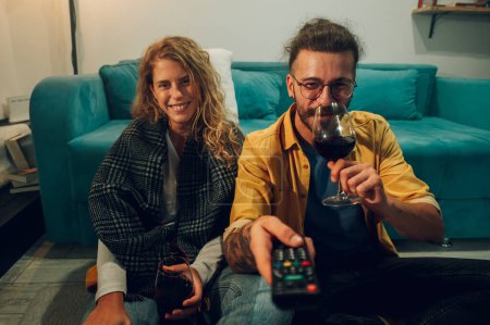 Front view shot of a happy caucasian couple sitting on the floor in front of the TV. Young man sipping red wine while changing the channel and his girlfriend sitting next to him and watching TV.