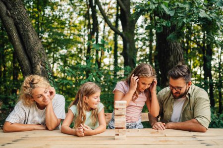 Eine fröhliche Familie sitzt am Tisch beim Picknick und spielt ein lustiges Lehrspiel in der Natur. Ein Mädchen summiert Zahlen auf Würfeln und plant ihre Strategie. Eine Familie spielt lustiges Jenga-Spiel.