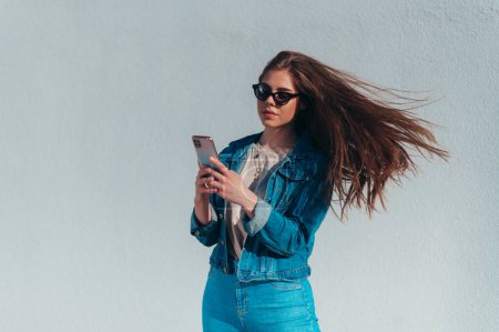 Junge schöne Frau mit Smartphone und Jeans und Sonnenbrille vor einer weißen Wand