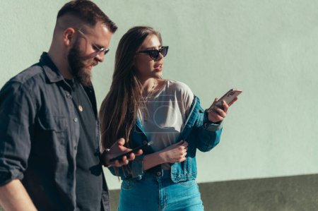 Atractiva pareja joven usando teléfonos inteligentes mientras está de pie frente a una pared blanca con gafas de sol
