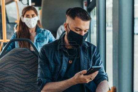 Mann mit Schutzmaske und Smartphone während der Busfahrt und auf Abstand zu anderen Passagieren wegen Covid19 Pandemie