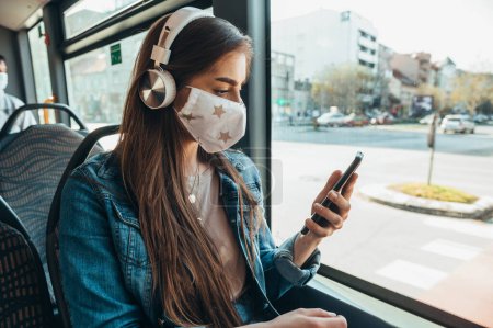 Junge schöne Frau mit Schutzmaske und Smartphone während der Busfahrt mit Kopfhörer auf dem Kopf