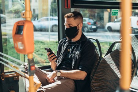Schöner Mann mit Schutzmaske und Smartphone während der Busfahrt und auf Abstand zu anderen Fahrgästen