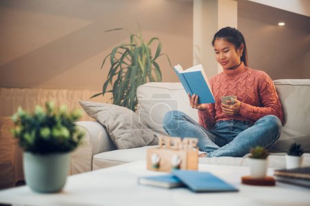 Glückliche junge asiatische Frau liest ein Buch, während sie zu Hause auf einem Sofa sitzt und Kaffee oder Tee trinkt. Lifestyle und Relax Konzept. Freizeit am Wochenende zu Hause genießen. Kopierraum.
