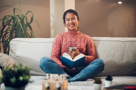 Junge entspannte und glückliche asiatische Frau liest ein Buch und trinkt Kaffee oder Tee, während sie es sich auf dem Sofa im gemütlichen Wohnzimmer gemütlich macht. Lässiger Lebensstil zu Hause. Blick in die Kamera. Bücherwurm. Kopierraum.