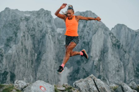 An extreme female skyrunner is running and jumping on rocky terrain in mountain range. An athlete is mountaineering and trail running while jumping on rocky terrain. In background is mountain range.