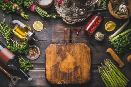 Food background. Various ingredients and seasonings for tasty cooking around  cutting board on wooden rustic table. Oil,lemon, vine bottle, pickled chili in jar, asparagus, herbs and spices. Frame