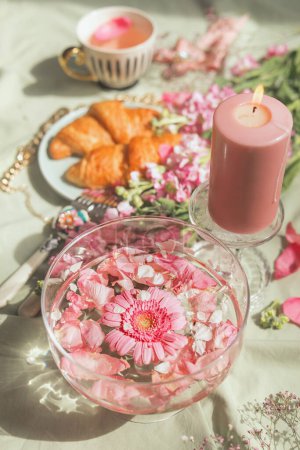 Aesthetic picnic breakfast with pink flowers in water bowl , candle, croissants and tea cup. Elegant food and drink concept. Fancy drink with flowers. Top view.