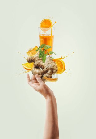 Women hand holding balancing ginger and orange immunity boosting drink with splashing and ingredients at light background. Creative healthy lifestyle. Levitation food . Front view