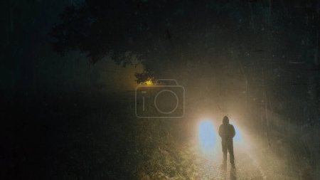 Foto de Mirando hacia abajo a una misteriosa figura encapuchada. De pie frente a los faros del coche. En una espeluznante noche de niebla en el campo. - Imagen libre de derechos