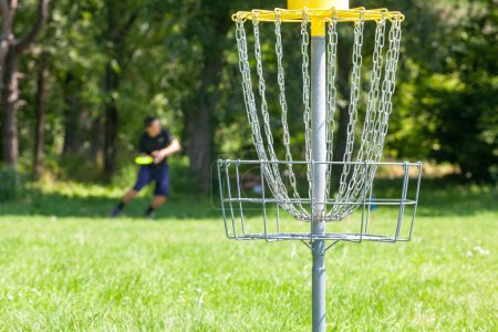 Téléchargez les photos : Joueur de golf de disque jetant un disque volant dans le parc, panier à chaîne au centre - en image libre de droit