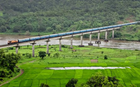 Téléchargez les photos : Le train super rapide traverse le long viaduc sur la rivière à l'emplacement pittoresque des champs verts sur la route ferroviaire Konkan. - en image libre de droit