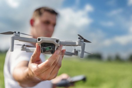 A quadcopter operator with a remote control out of focus holds a drone in front of him in focus outdoor.