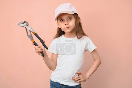 Photo for Caucasian girl of primary school age in a baseball cap with a wrench on a pink background. - Royalty Free Image