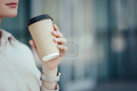 Close-up side view of unrecognizable young business woman holding in hands paper cup with takeaway coffee or other beverage.