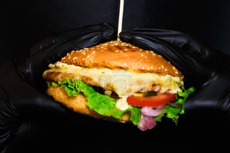 Téléchargez les photos : A close-up view on a cheeseburger holding in the hands dressed in black glooves. On a black background - en image libre de droit