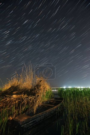 Téléchargez les photos : Night cosmic star trails landscape with boat floating on a river's water surface. Long exposure rural photography - en image libre de droit