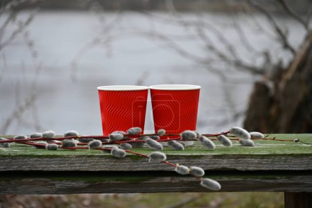 Téléchargez les photos : Deux tasses en papier rouge remplies de café aromatique se tiennent sur un banc en bois entouré de branches de saule dans le contexte d'une rivière calme. - en image libre de droit