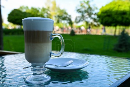 a large glass with aromatic, delicious latte coffee on the table of a street cafe in a park where people are walking on a warm sunny day.